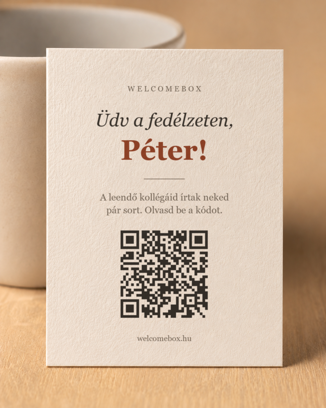 Névre szóló krémszínű kártya: „Üdv a fedélzeten, Péter!” felirat és egy QR-kód, egy bögrének támasztva.